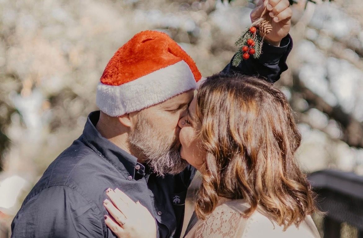 Couple sharing a Christmas kiss under mistletoe, man wearing a Santa hat outdoors in winter.