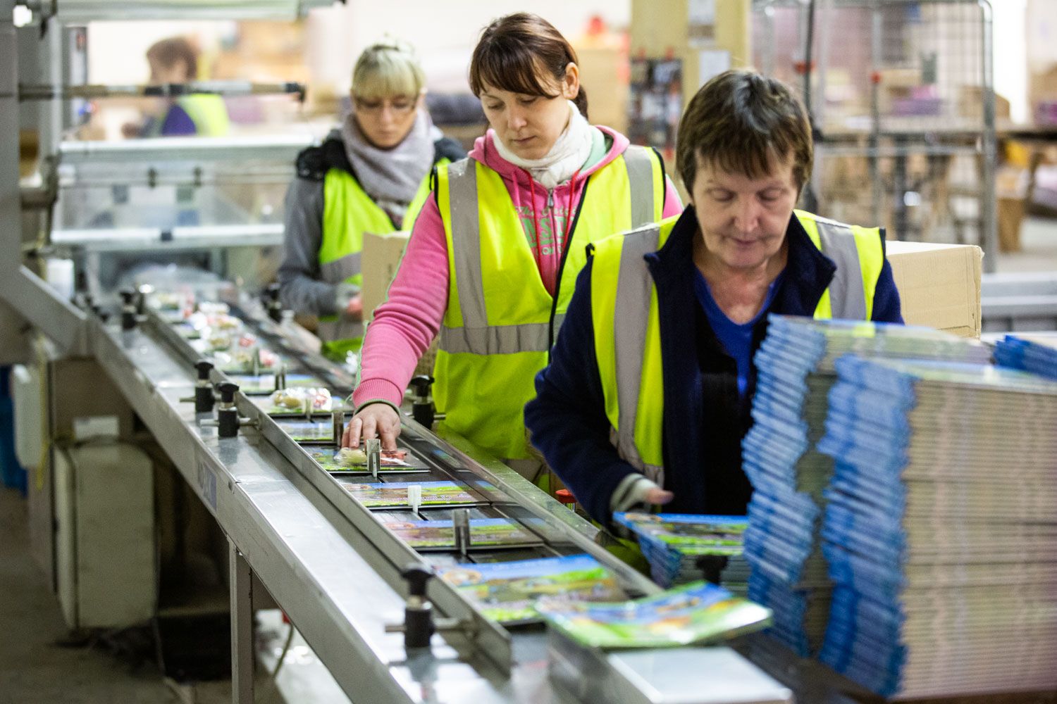 Workers in yellow safety vests sort books on a conveyor belt in a busy warehouse fulfillment center.