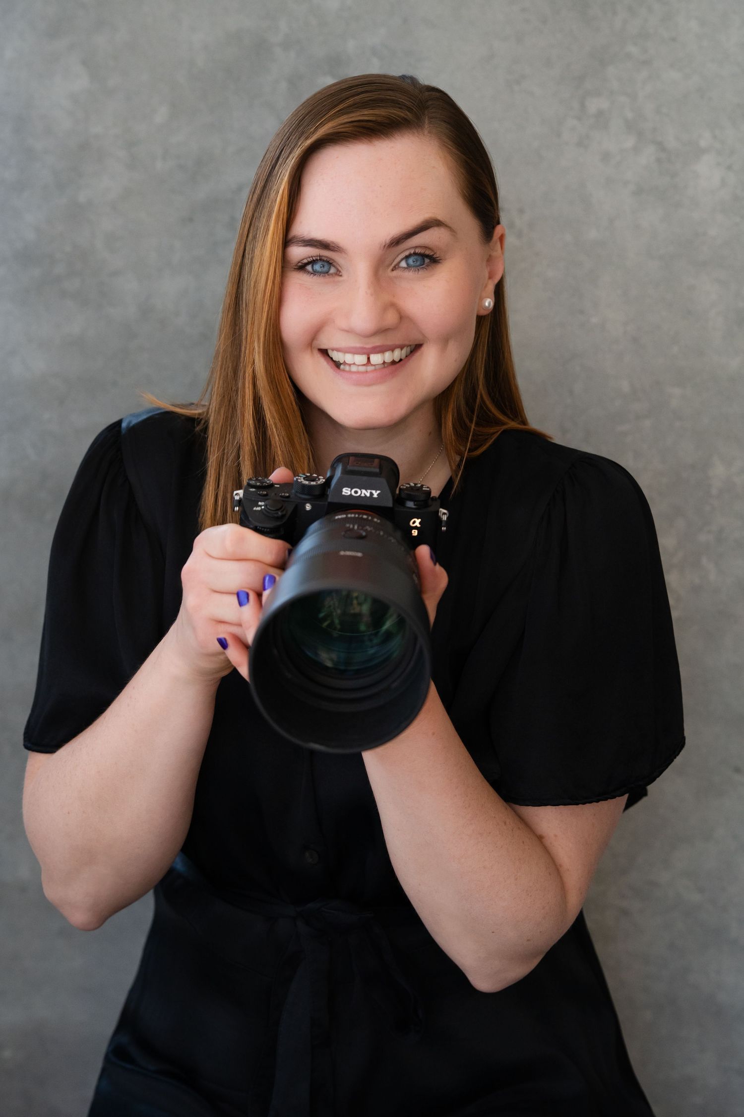 Smiling female photographer holding a Sony camera, wearing black top, with long brown hair and blue eyes.