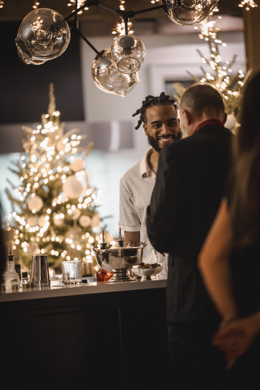 Smiling bartender serves guests at a festive holiday party with decorated Christmas trees and warm string lights.