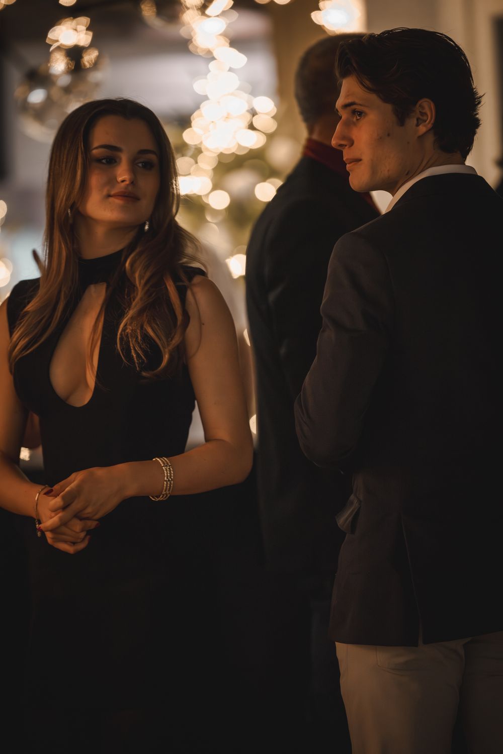 A young woman in a black dress and a man in a suit stand together at an elegant evening event with bokeh lights.