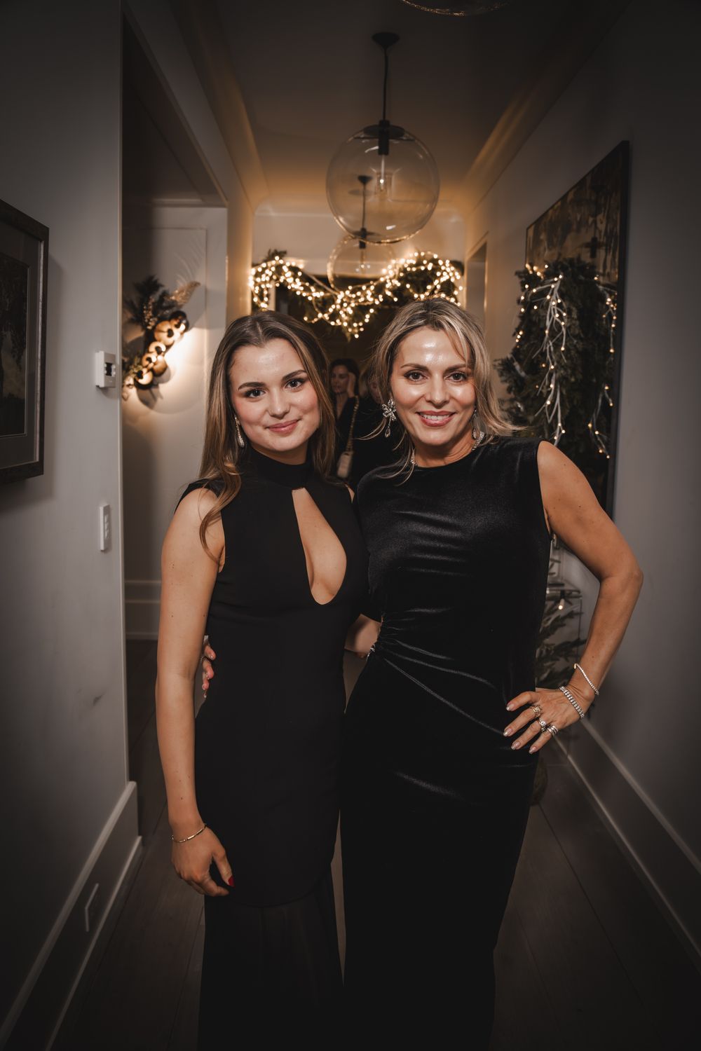 Two women in elegant black dresses posing together in a decorated hallway at a festive evening event.