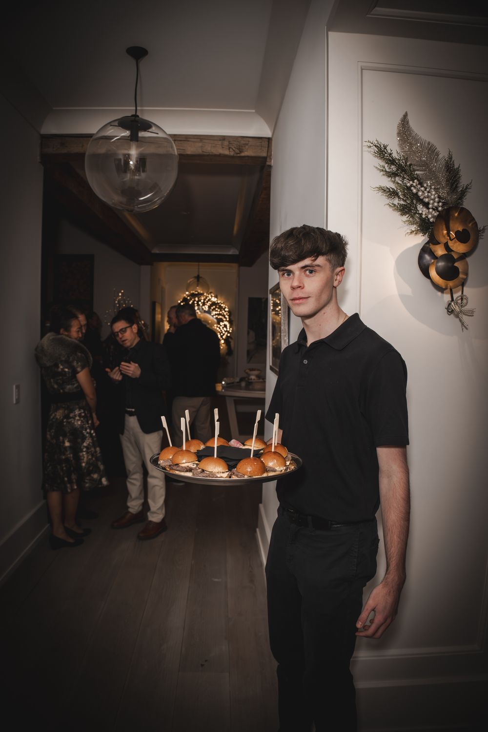 Young waiter in black uniform holding a tray of gourmet sliders with candles at an elegant indoor party.