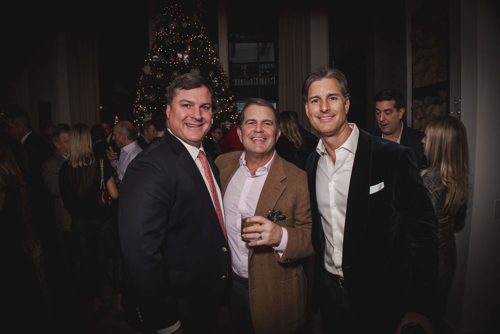 Three men smiling together at a festive holiday party with a decorated Christmas tree in the background.