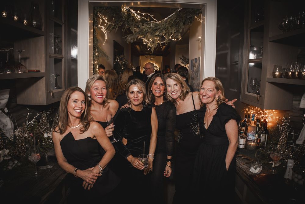 Six women in black dresses pose together smiling at an elegant evening party with warm ambient lighting.