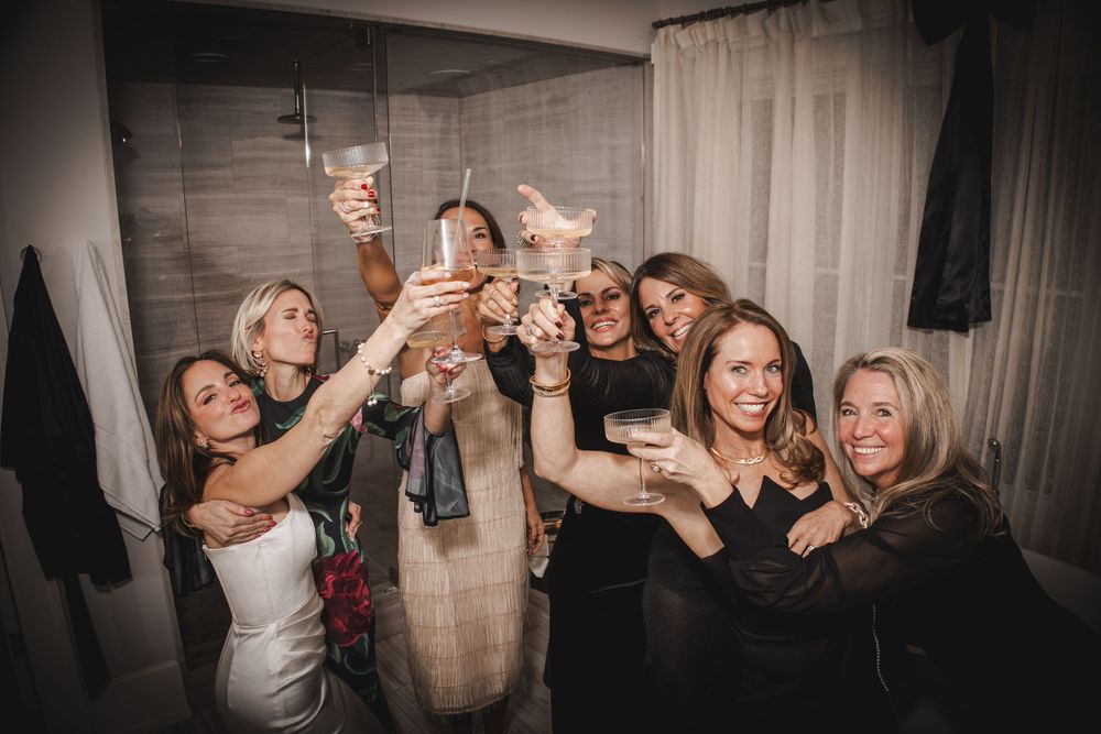 Six women in elegant attire raising glasses in a celebratory toast at a stylish indoor party.