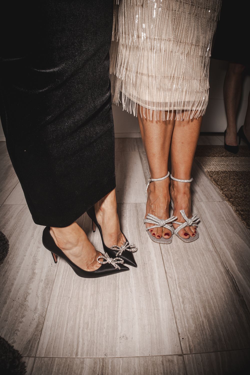 Two women standing together, one in black heels with bow detail, other in strappy silver sandals, wearing elegant formal attire.