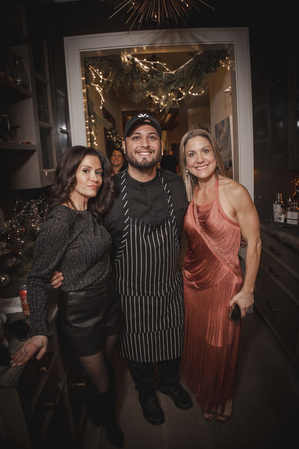 Chef in striped apron poses with two women in festive attire at a holiday restaurant event.