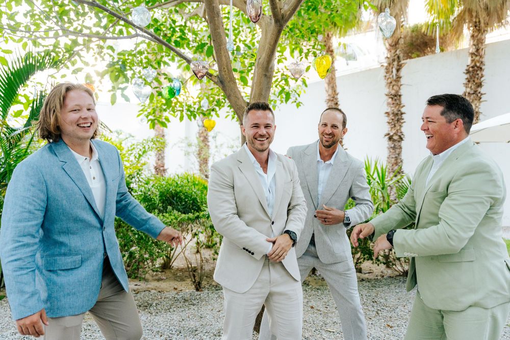 Four men in light-colored suits laughing and celebrating outdoors near a tree with colorful decorations.