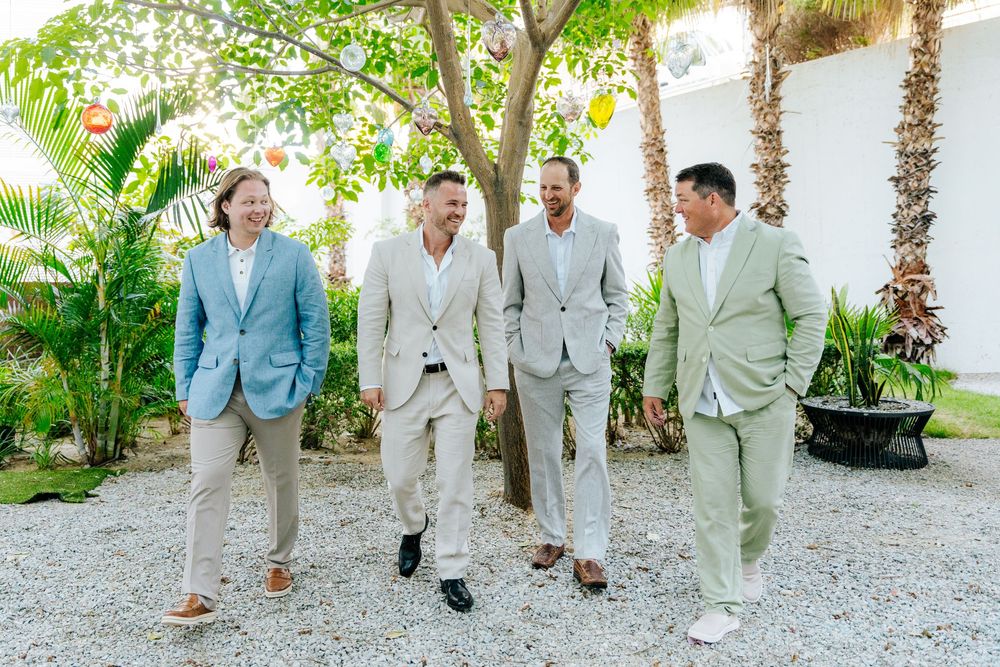 Four groomsmen in linen suits laughing and walking together outdoors at a tropical wedding venue.