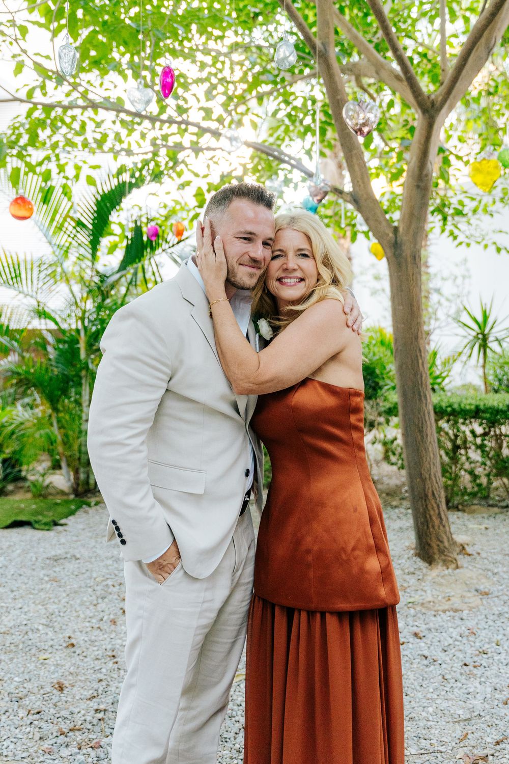 Couple embracing outdoors under a tree with colorful decorations, man in beige suit and woman in rust satin dress.