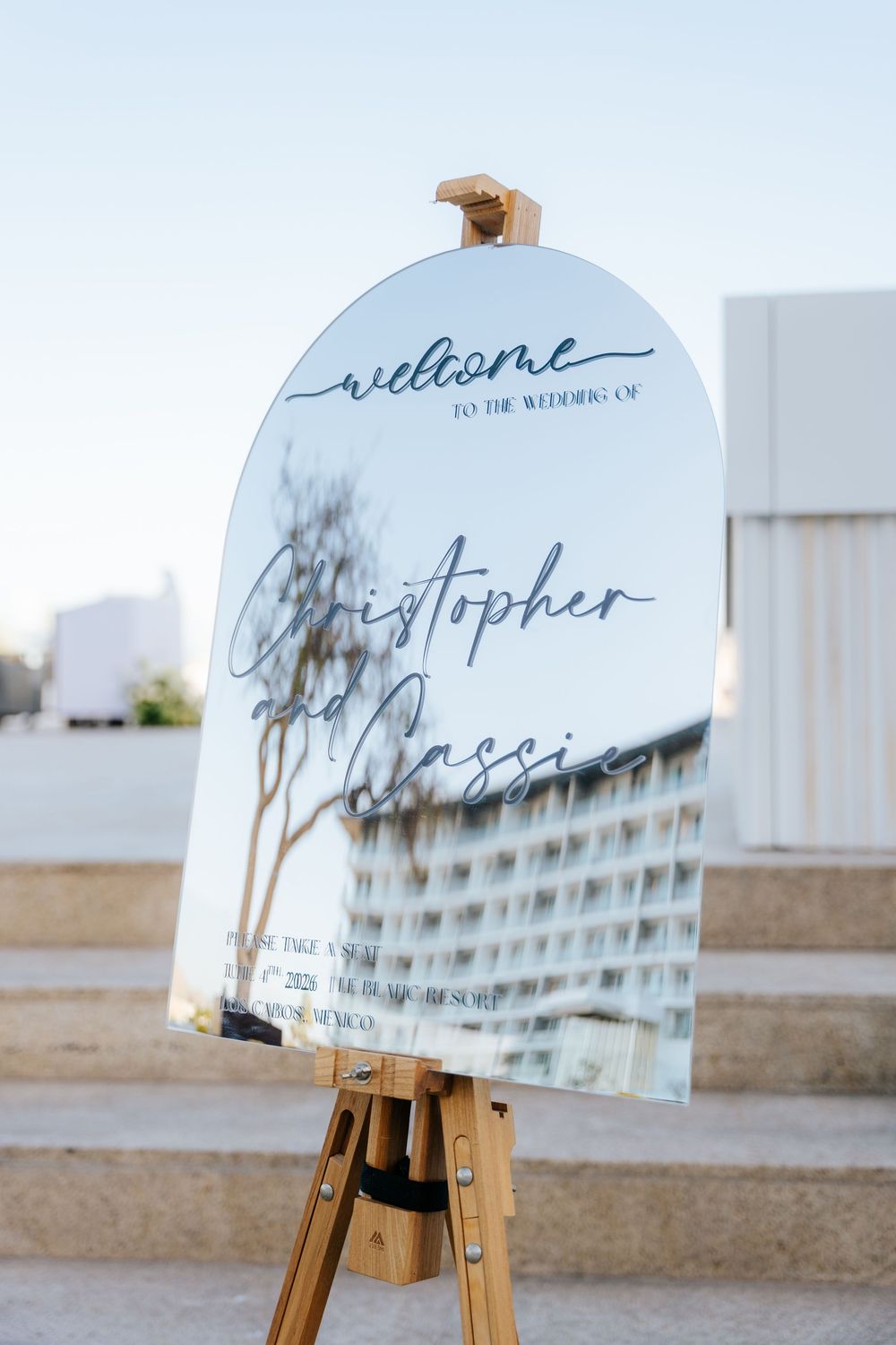 Elegant mirrored wedding welcome sign on wooden easel displaying Christopher and Cassie's wedding details outdoors.