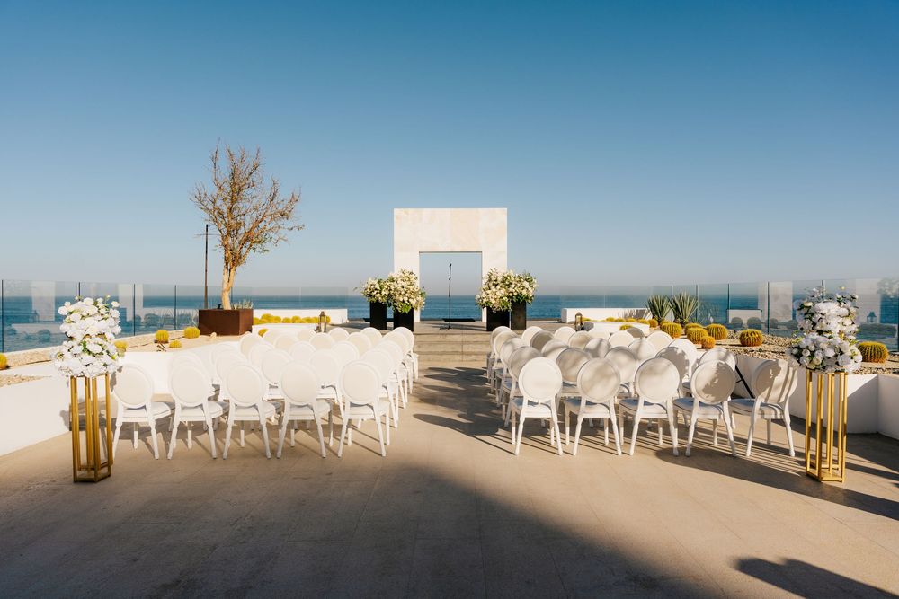 Elegant rooftop wedding ceremony setup with white chairs, gold accents, floral arrangements, and ocean views under a clear blue sky.