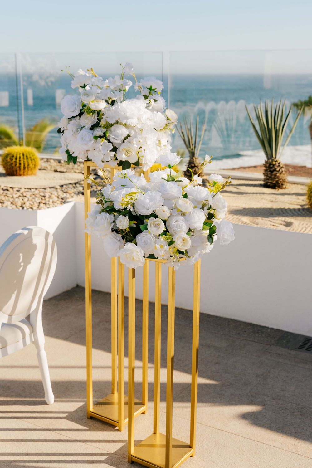 Gold pedestal stands with white floral arrangements at an outdoor wedding venue overlooking the ocean.