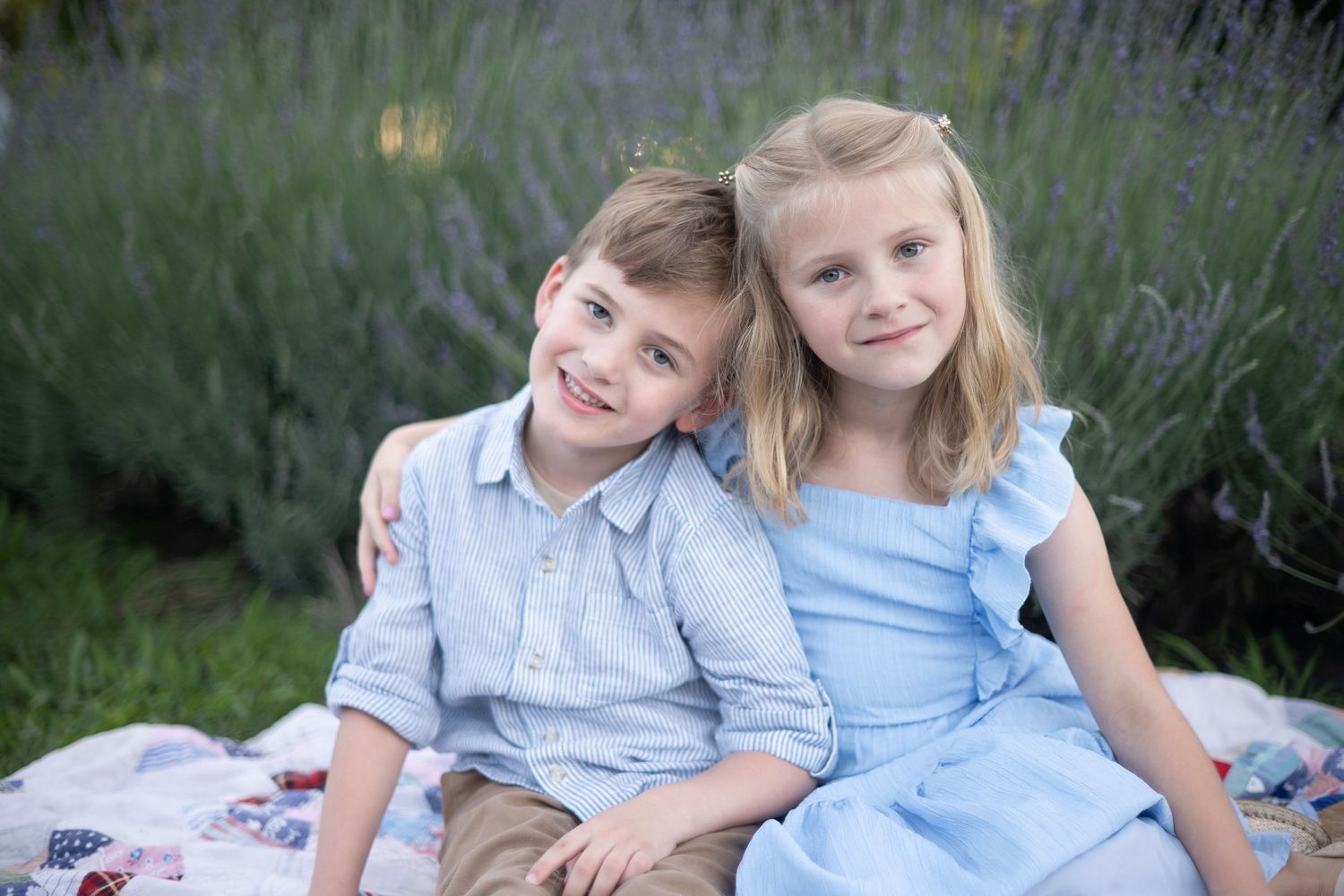 Two smiling children in light blue outfits hugging outdoors with purple lavender flowers in the background.