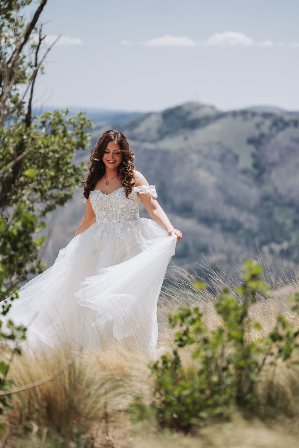 Smiling bride in white lace off-shoulder gown twirling outdoors with mountain landscape backdrop on sunny day.