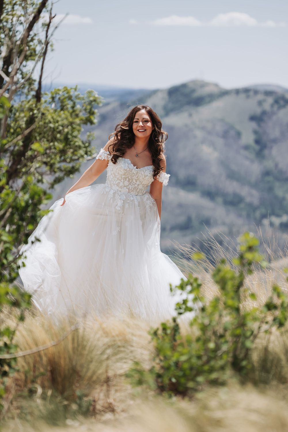 Smiling bride in off-shoulder lace wedding gown holding skirt in mountain landscape.