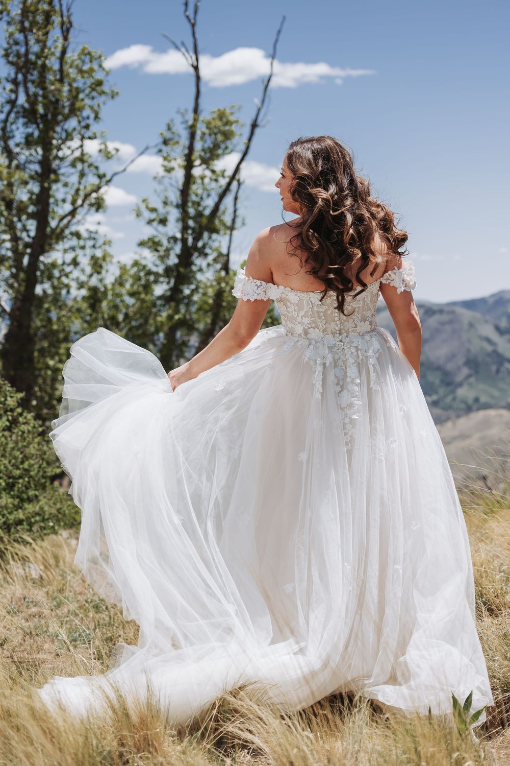 Bride in flowing off-shoulder lace gown viewed from behind in scenic mountain setting.