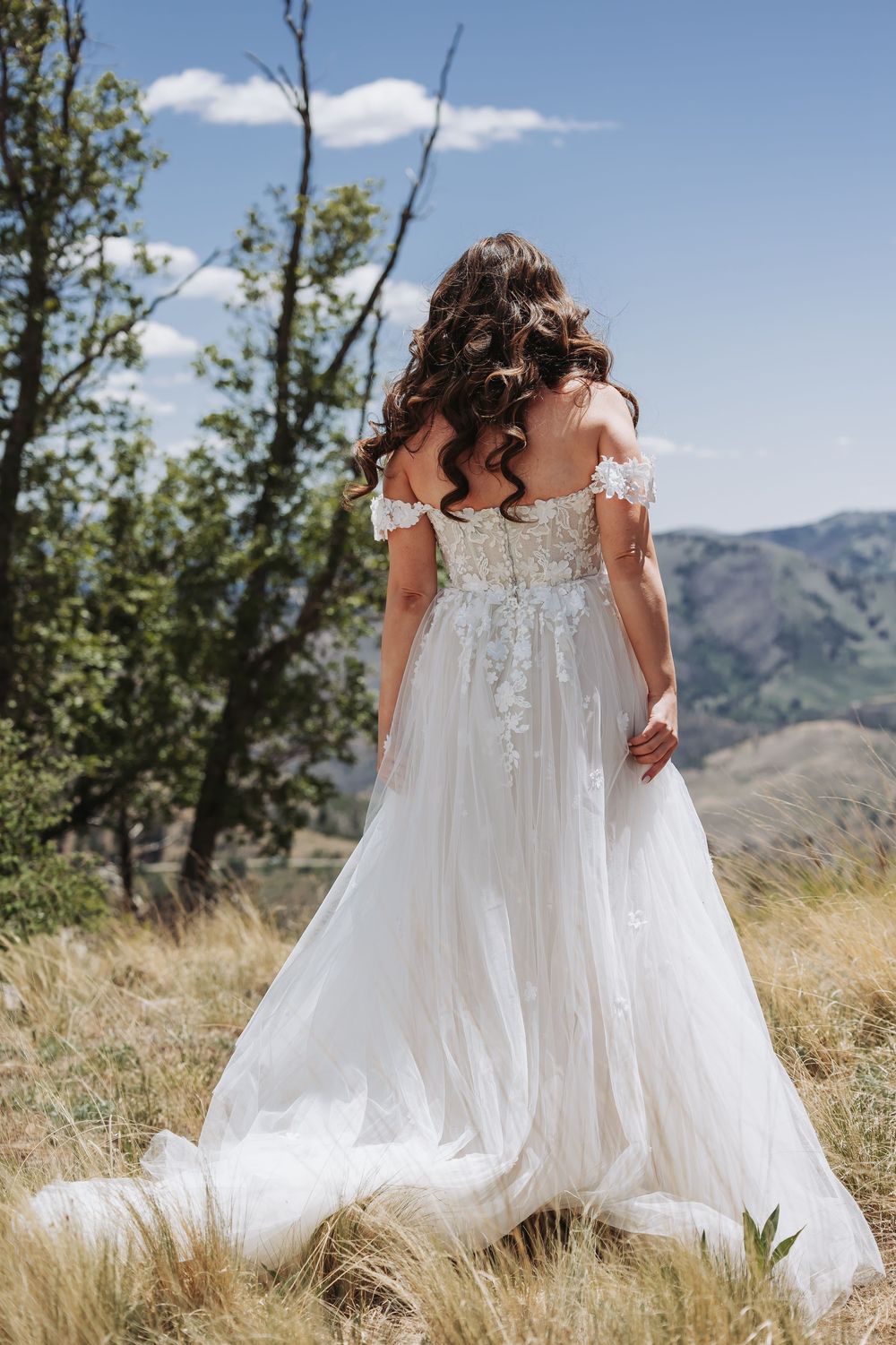 Bride in white off-shoulder lace wedding gown with train stands in mountain meadow, back view, curly brunette hair.