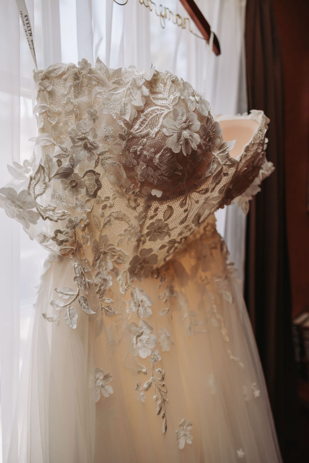 Elegant strapless wedding gown with intricate floral lace appliques and flowing tulle skirt hanging by a window.