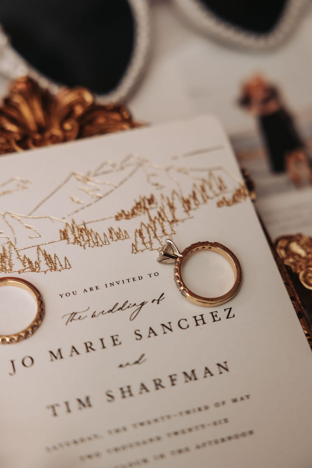 Gold wedding rings rest on an elegant wedding invitation for Jo Marie Sanchez and Tim Sharfman with mountain illustration.