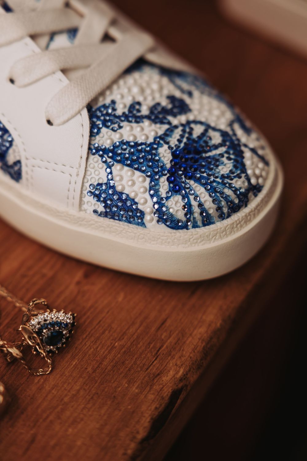 White sneaker adorned with blue crystal and pearl floral embellishments displayed on a wooden surface.