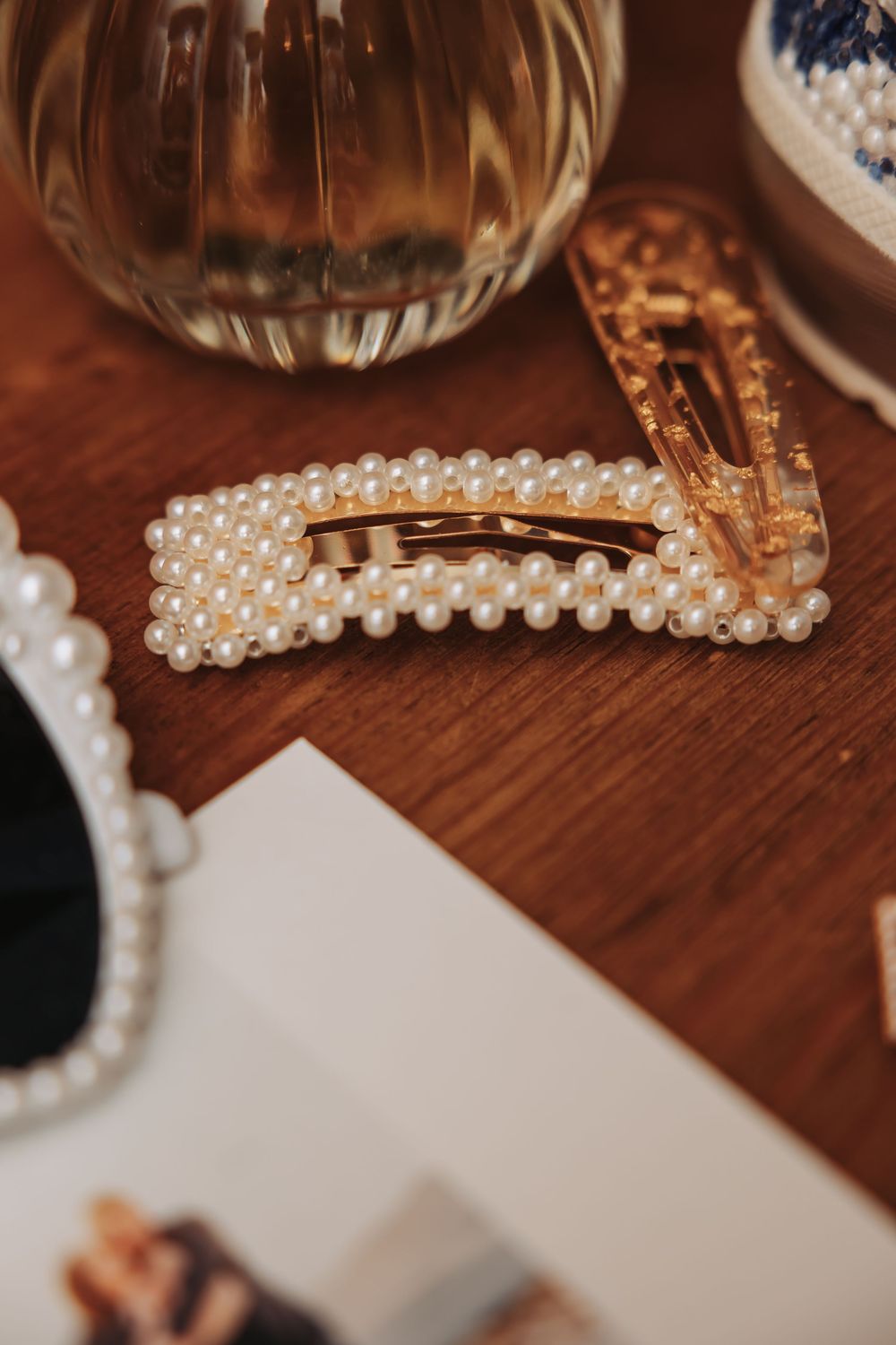 Gold hair clip adorned with pearl beads on a wooden surface, with a decorative mirror and glass vase in the background.