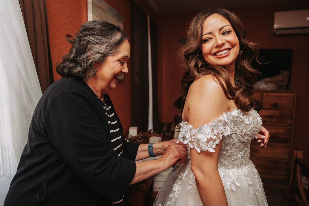 Older woman helping a smiling bride button her floral off-shoulder wedding gown indoors.