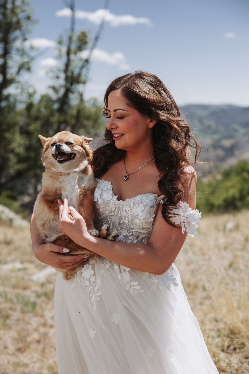 Bride in white lace off-shoulder gown holds smiling Corgi dog outdoors with mountain landscape background.