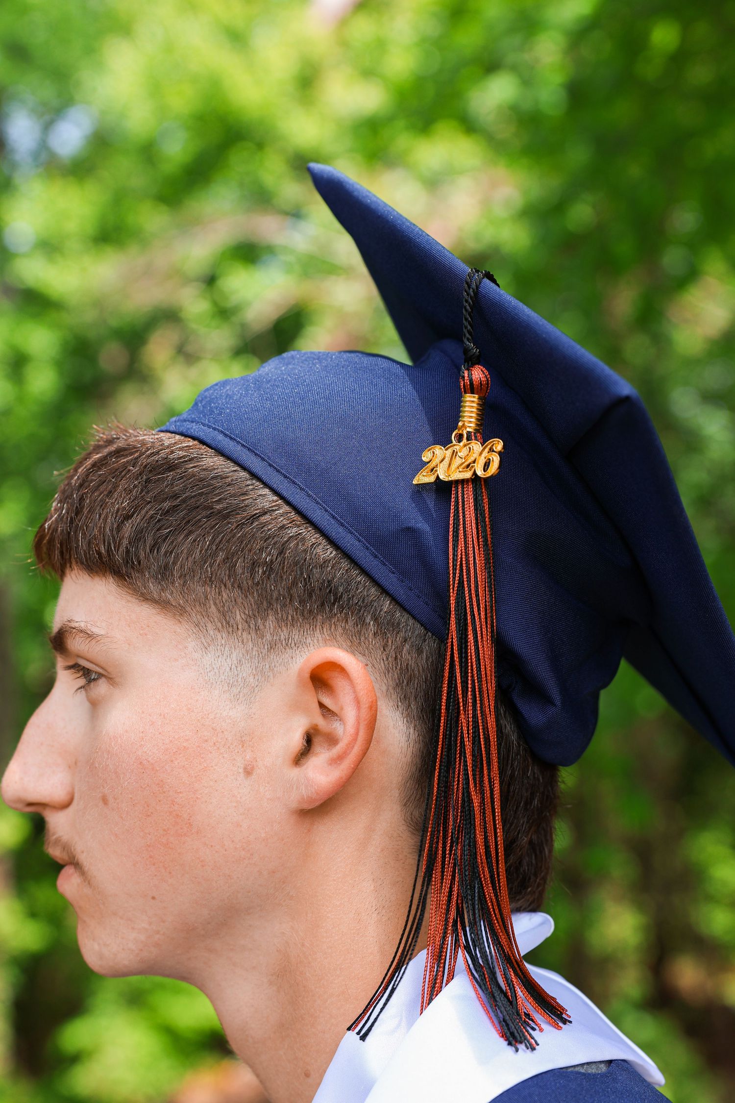 Young man in profile wearing navy blue 2026 graduation cap with orange and black tassel outdoors.