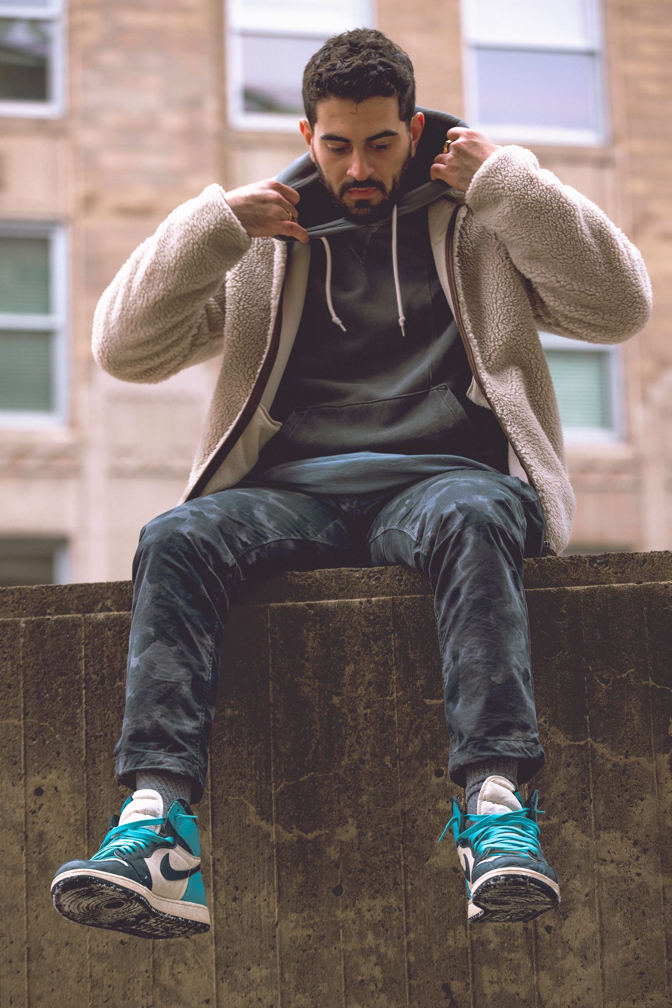 Bearded photographer crouching with camera, wearing gray hoodie, black jeans, and teal sneakers in modern corridor.