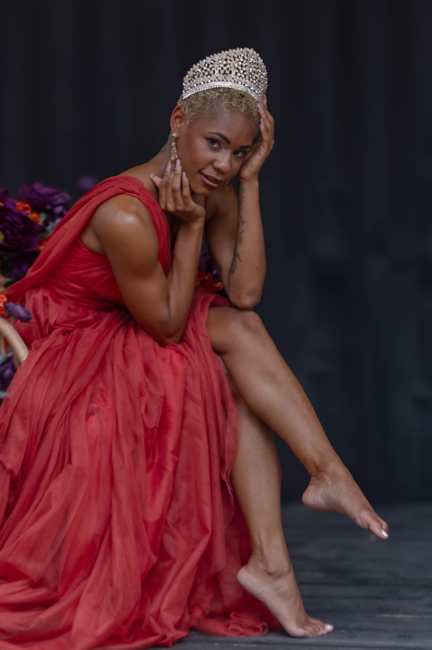 Woman in red gown and diamond tiara poses gracefully while seated, wearing elegant drop earrings against dark backdrop.