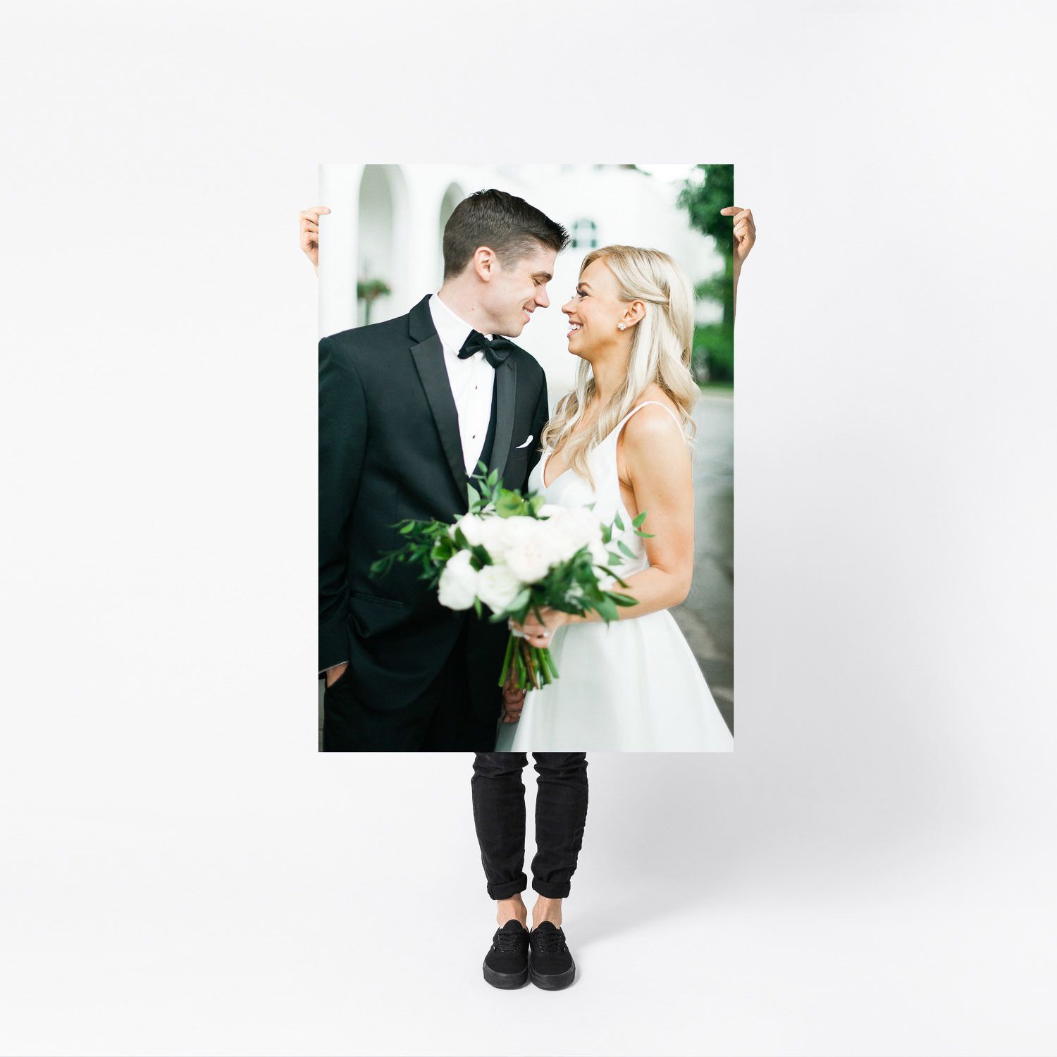 A person holds a large portrait print of a smiling bride and groom in wedding attire against a white background.
