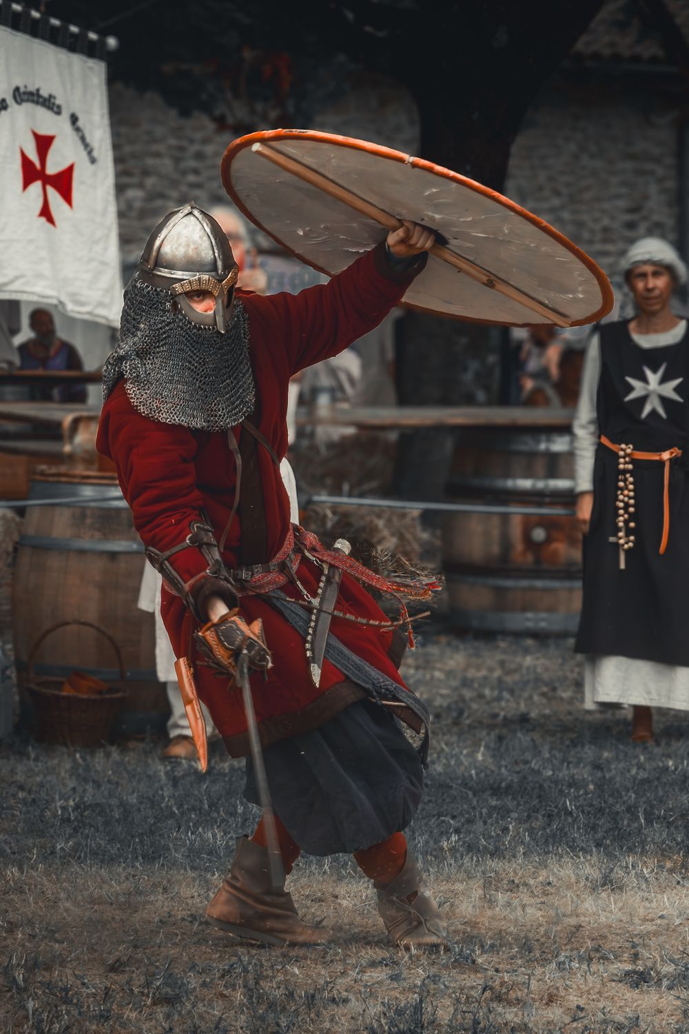 Medieval knight in chainmail and red surcoat wielding a shield and sword during a historical reenactment event.