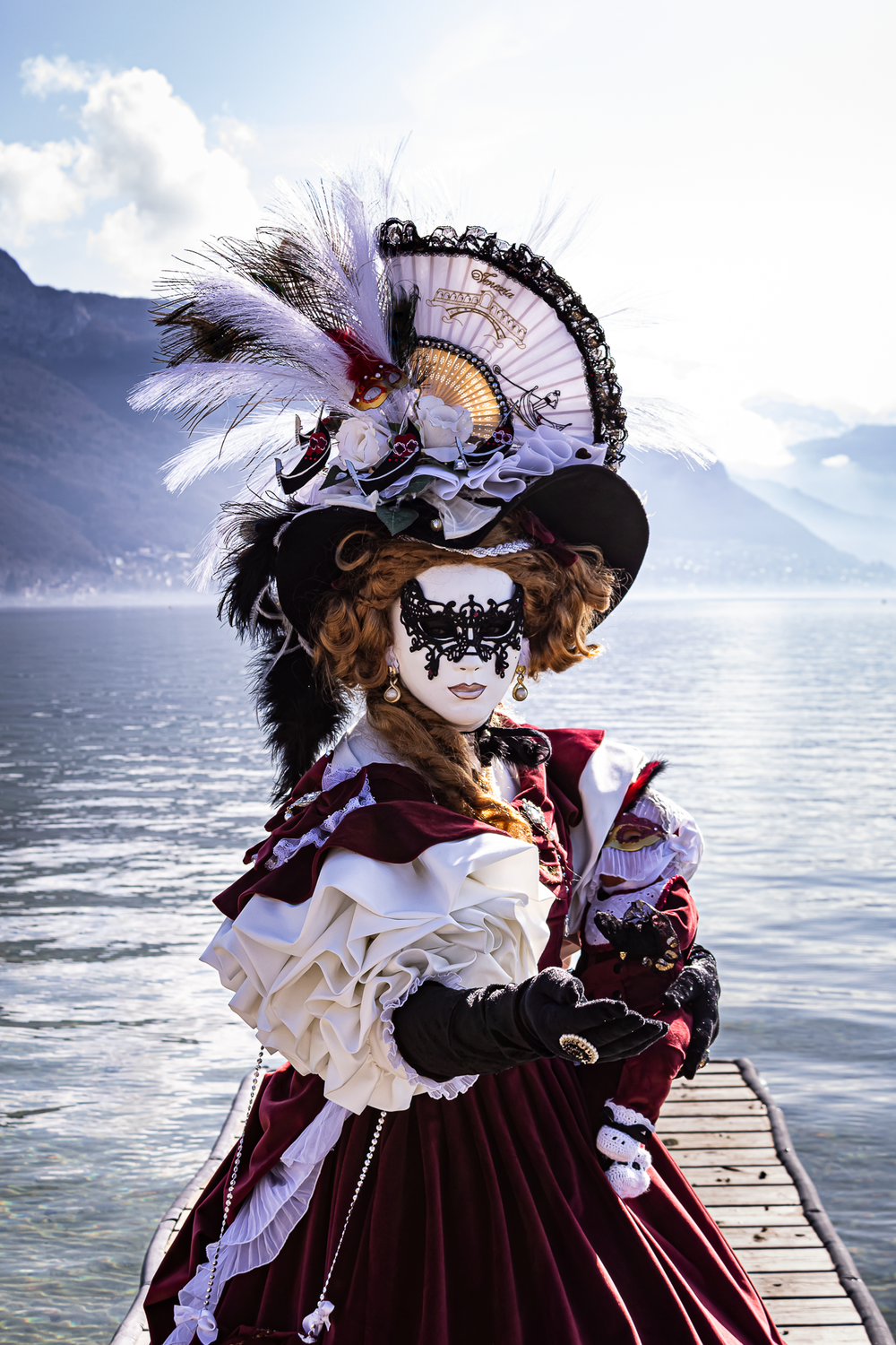 Elaborate Venetian carnival costume with ornate mask, feathered hat, and burgundy gown on a lakeside dock.