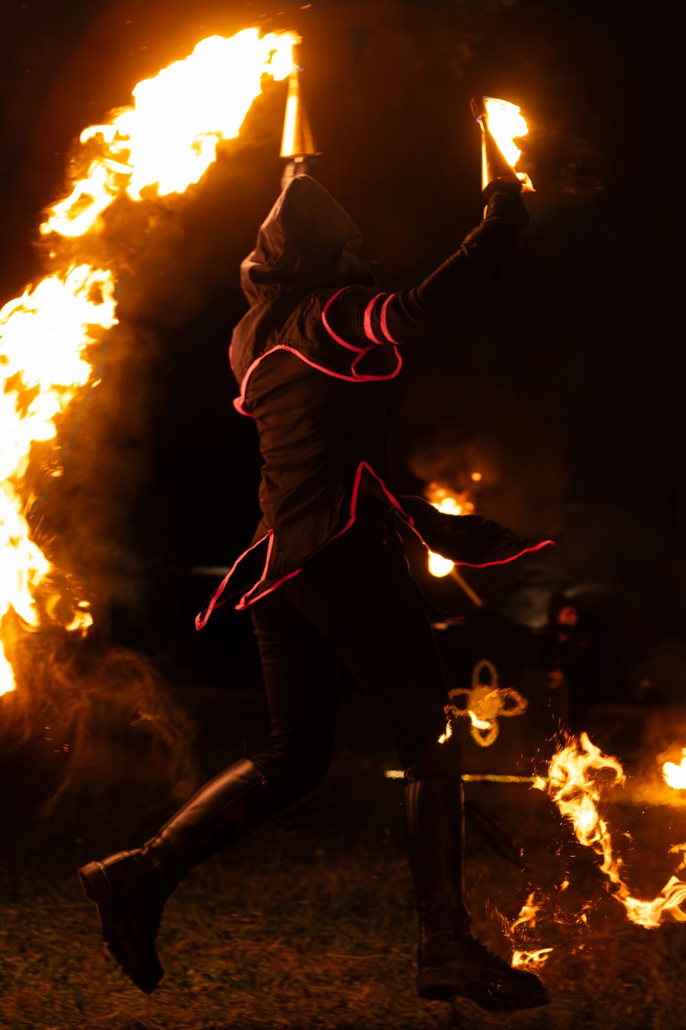 Fire performer jumping mid-air holding flaming torches at night, wearing hooded costume with glowing red accents.