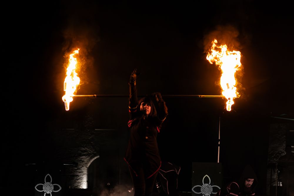 Fire performer holds flaming torch on dark stage with two large fire columns blazing dramatically.