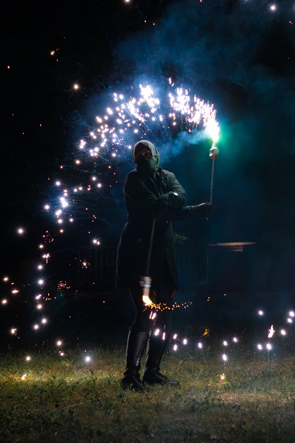 Person in dark costume holding sparkling firework torch at night with glowing embers surrounding them.