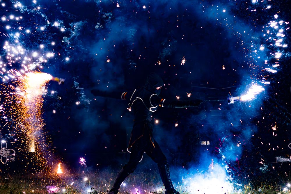 Silhouette of a performer surrounded by dazzling fireworks and sparks against a vivid blue night sky.