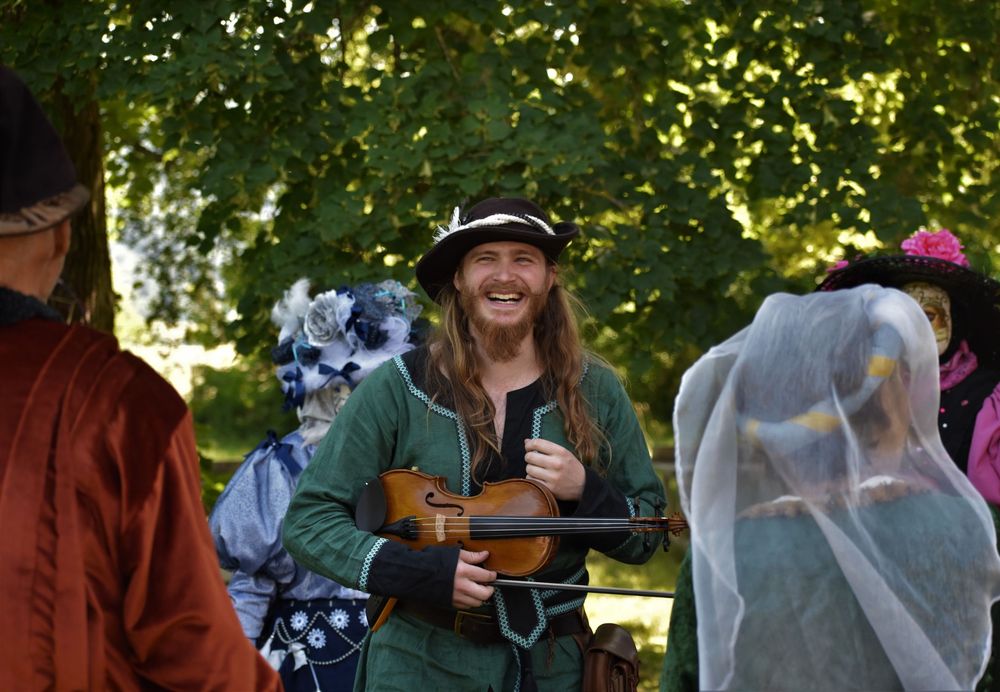 Smiling man with long hair holding a violin at a Renaissance faire, wearing a green costume and black hat.