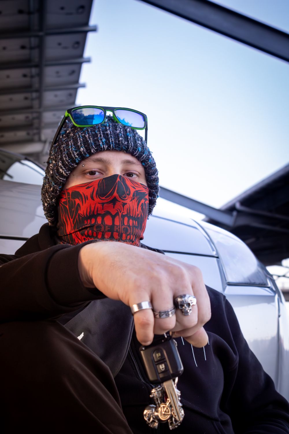 Person wearing red skull face mask, beanie, sunglasses, rings, holding car keys outdoors.