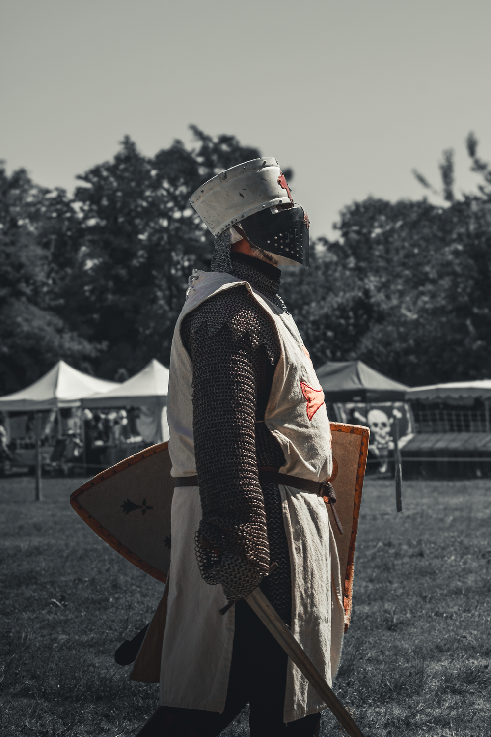 Medieval knight in chainmail armor and helmet carrying a shield at an outdoor historical reenactment event.