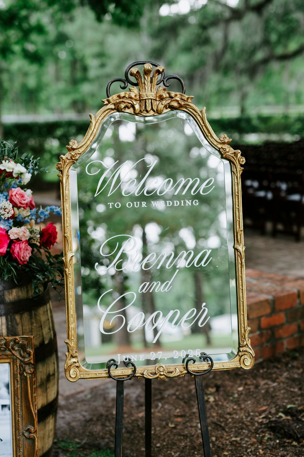 Gold ornate mirror wedding welcome sign for Brenna and Cooper displayed on easel outdoors amid lush greenery.
