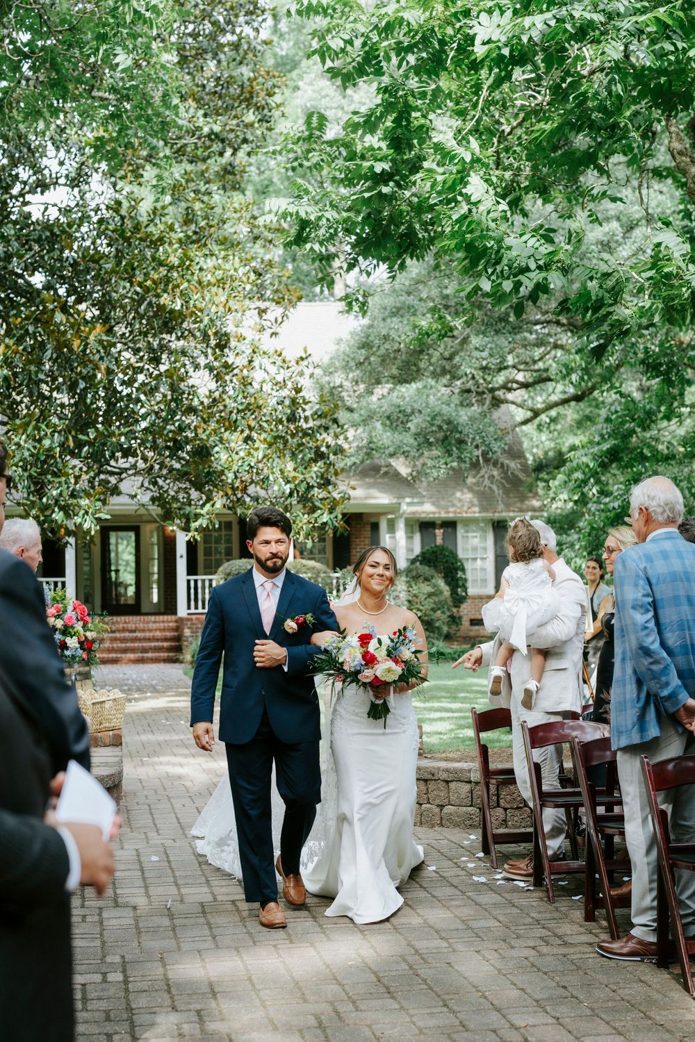 Bride and groom walk down outdoor wedding aisle lined with guests amid lush green trees.