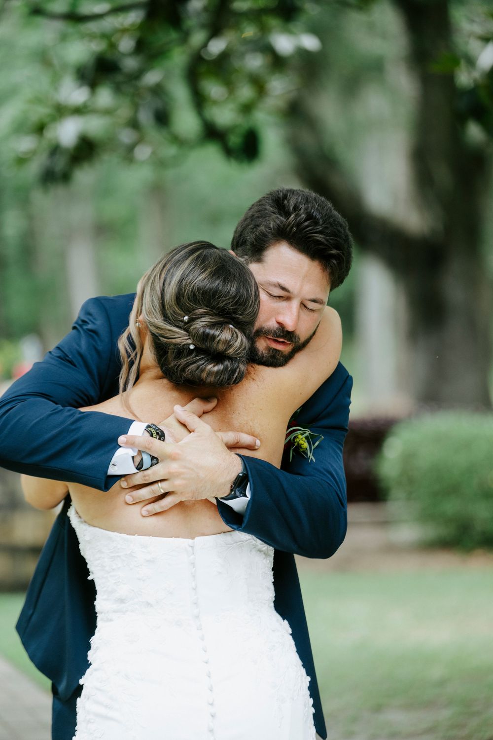 Groom in navy suit embracing bride in white strapless gown during emotional first look at outdoor wedding.