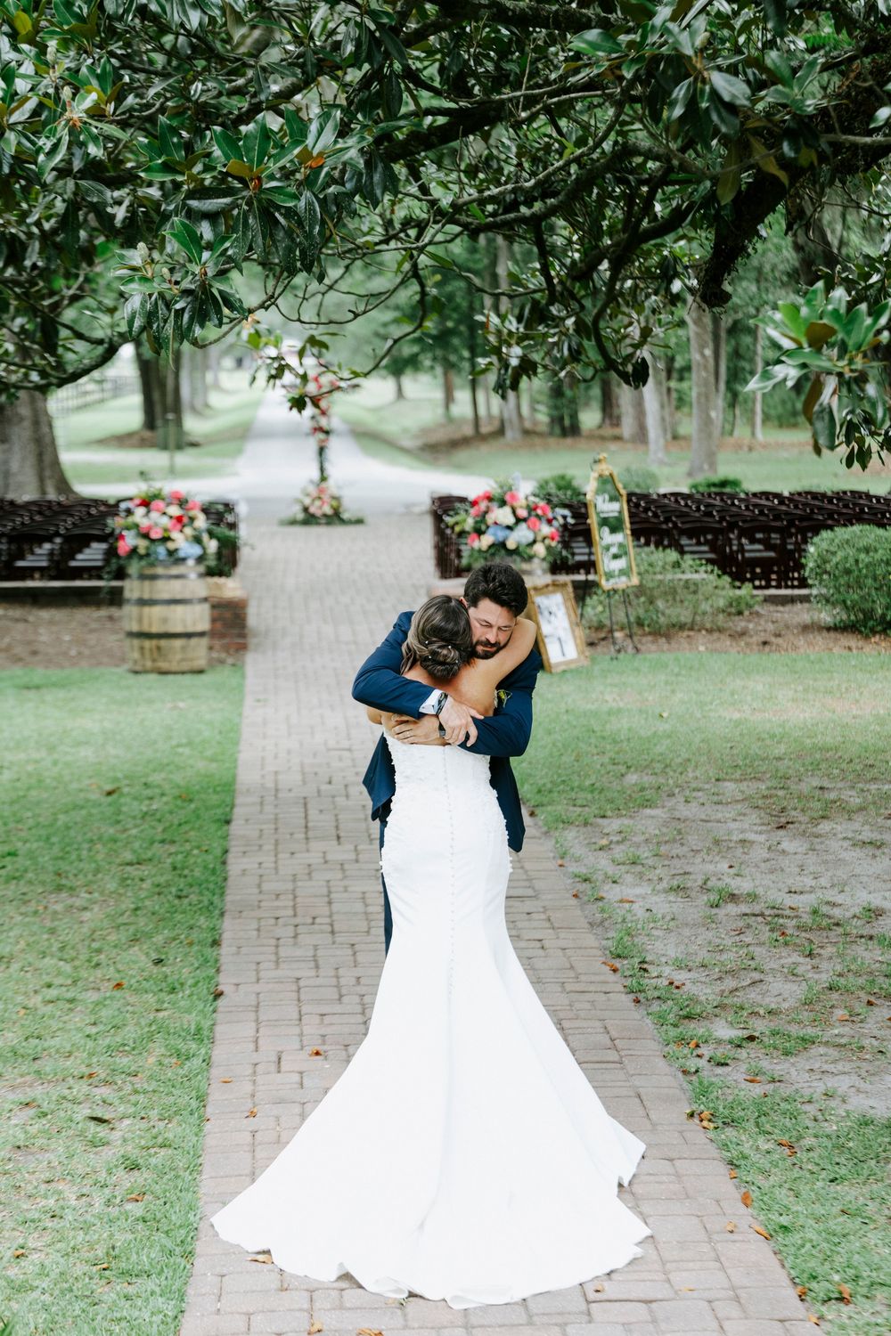 Bride and groom share a romantic embrace on a brick pathway lined with wooden barrels and floral arrangements at an outdoor wedding.