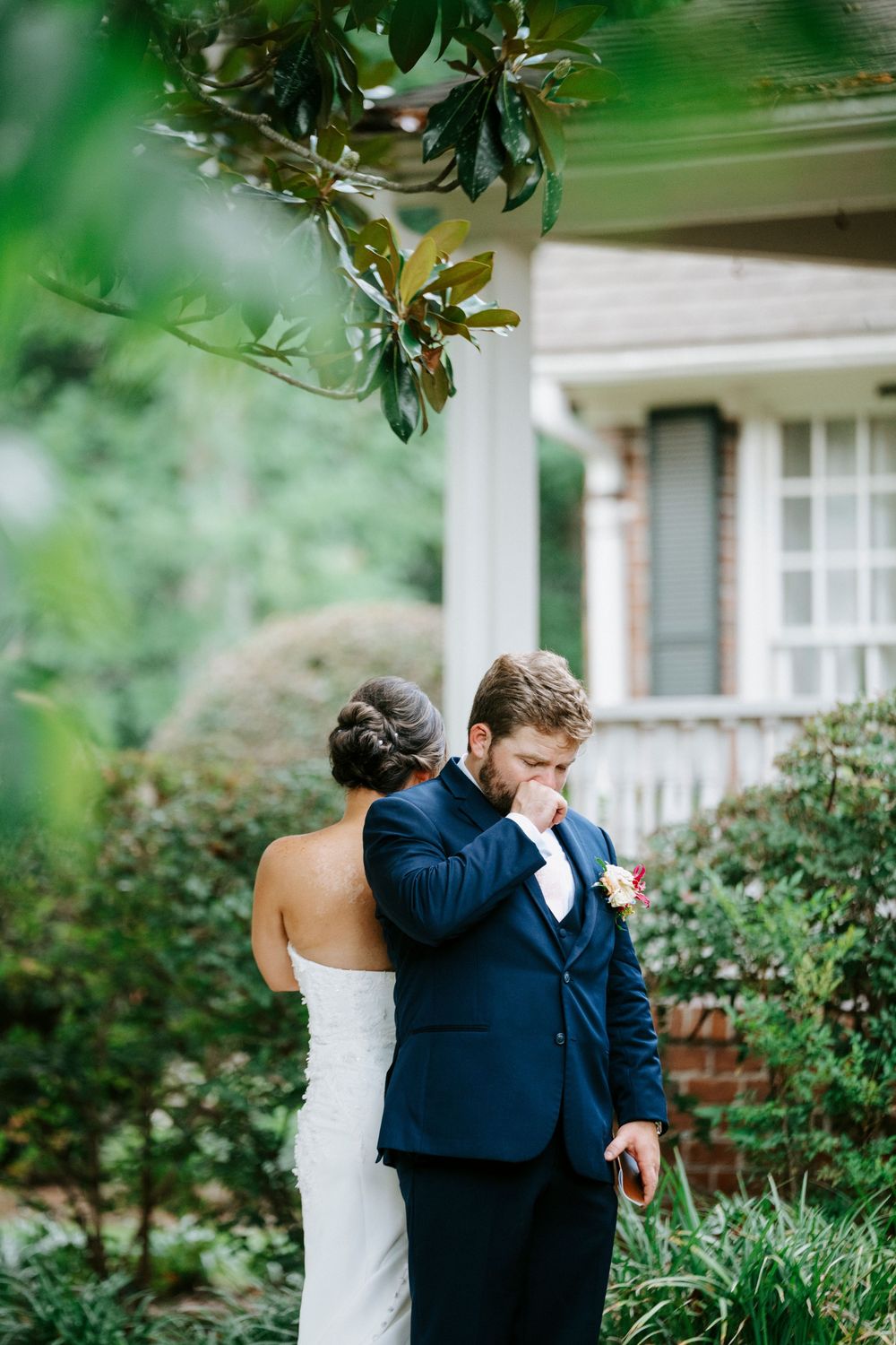 Emotional groom in navy suit wiping tears during first look with bride in white strapless gown outside brick home.