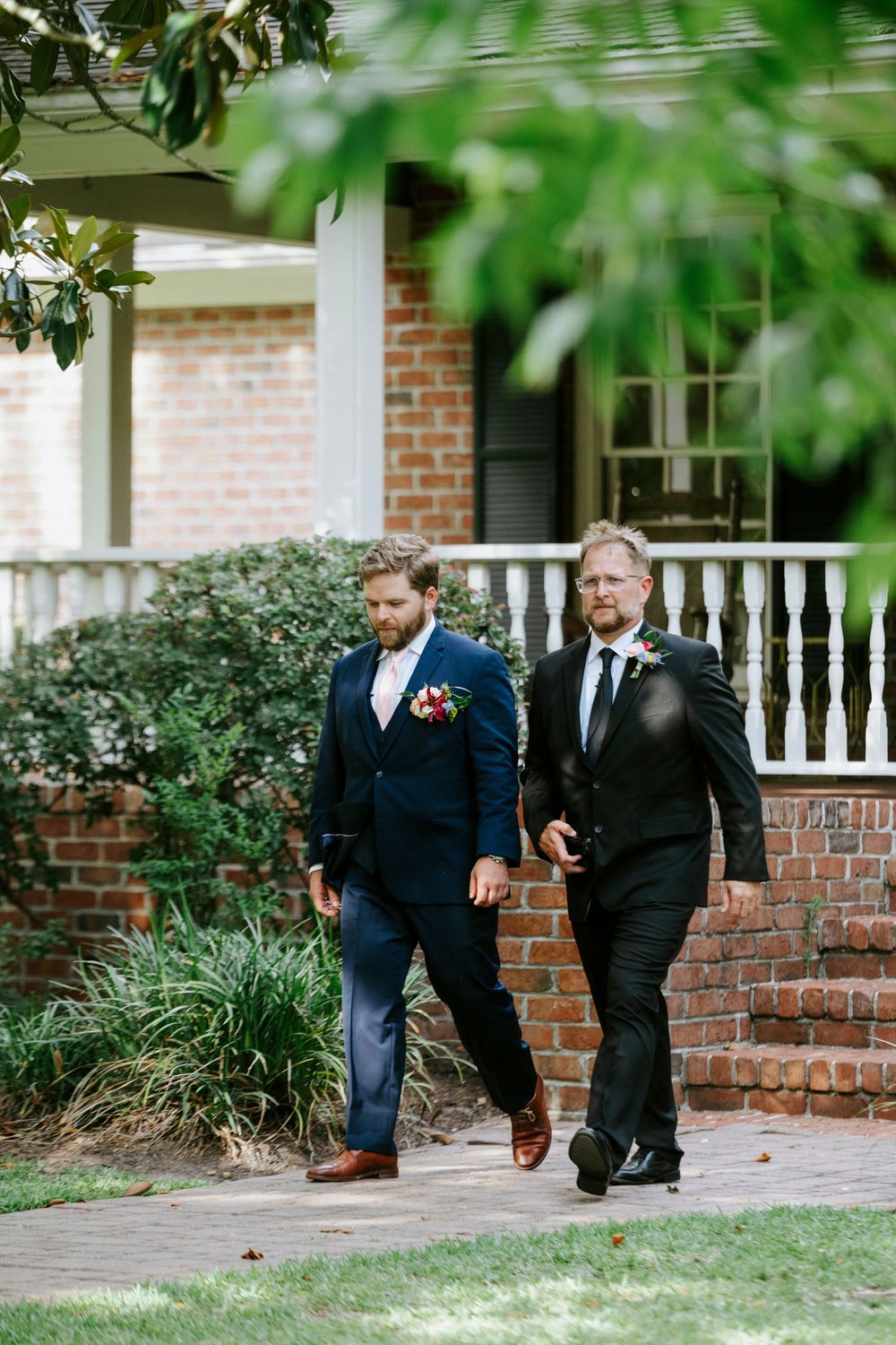 Two men in suits walking along brick path outside elegant venue during wedding celebration.