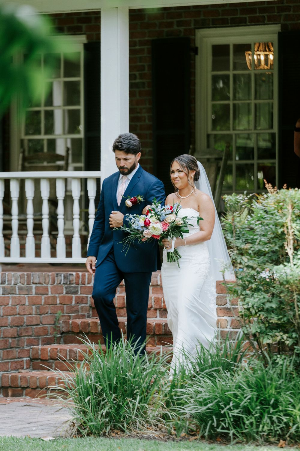 Bride in white gown with veil and groom in navy suit descend brick steps at elegant venue.