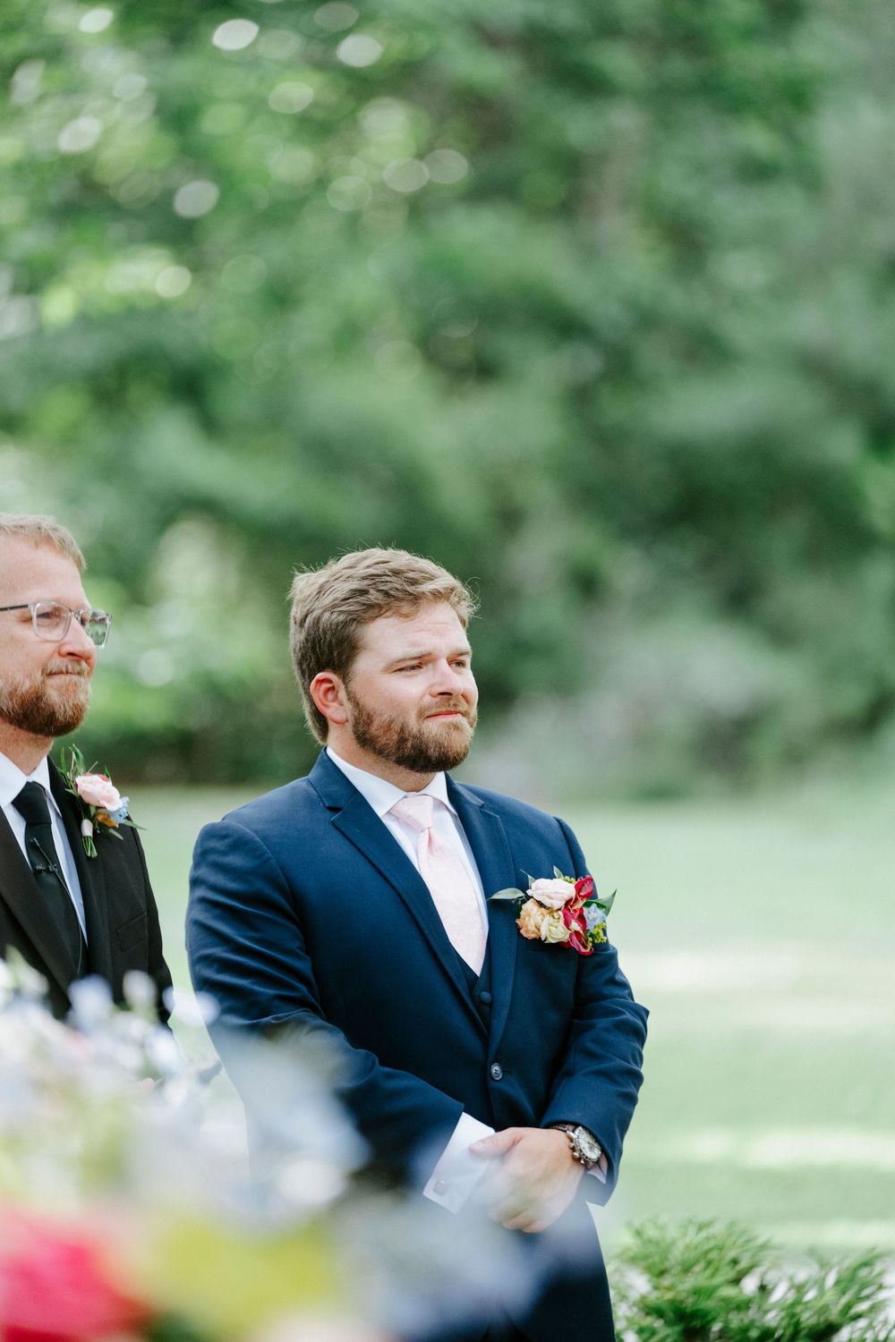 Groom in navy suit with floral boutonniere watches emotional ceremony at outdoor wedding.